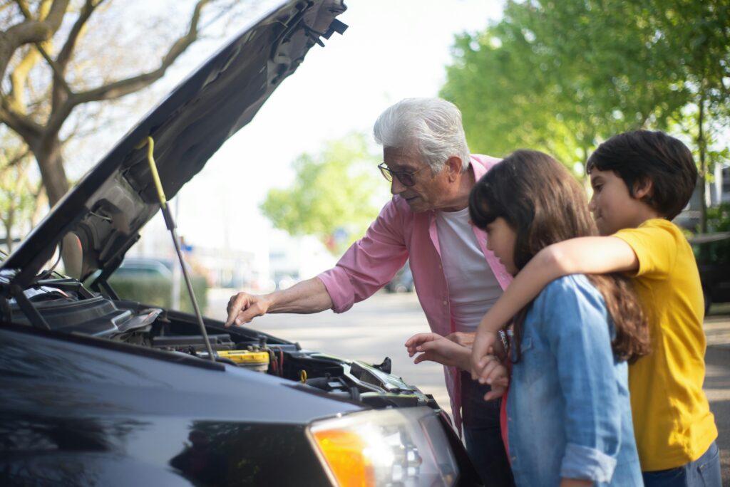 Grandfather shows his grandchildren car engine mechanics outdoors, teaching them about car maintenance.